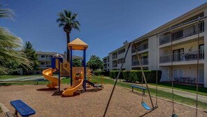 A playground with a slide and swings, surrounded by a building with balconies.