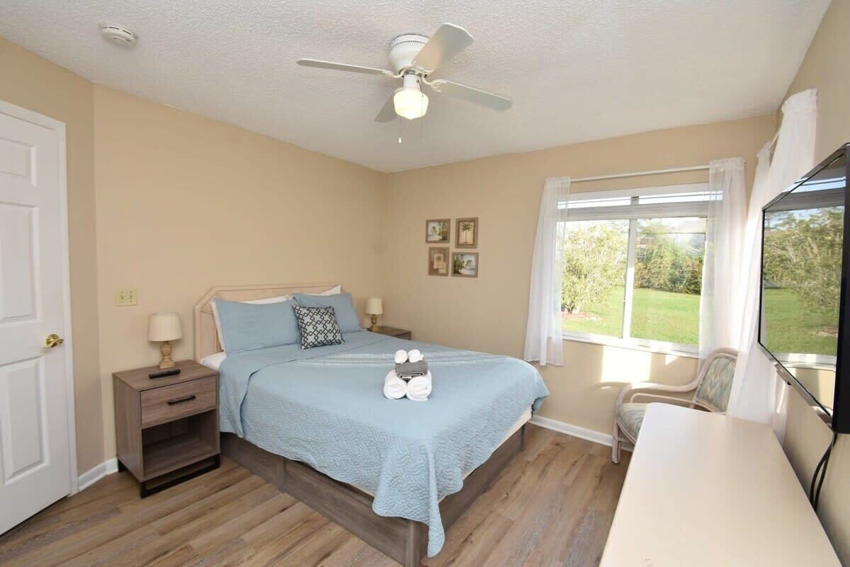 A bedroom with a bed, bedside tables, a ceiling fan, a window with curtains, and a view of greenery outside.
