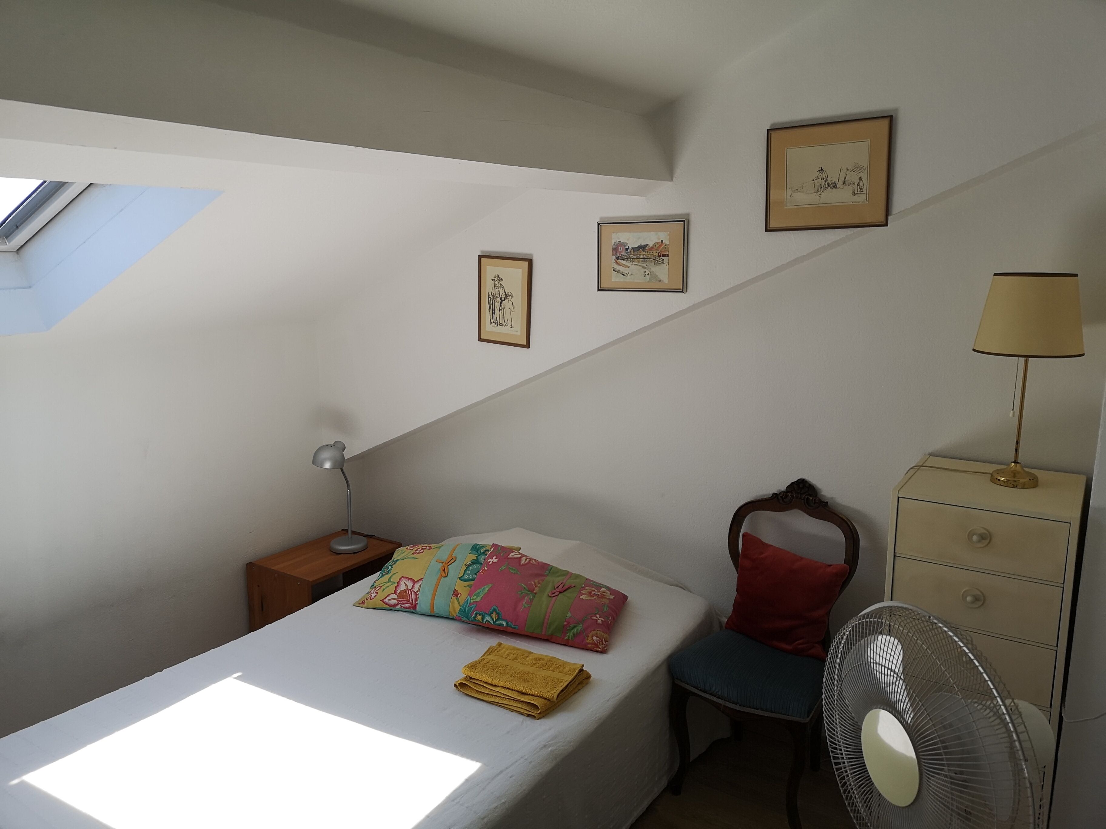 A bedroom with a bed, a nightstand, a chair, a dresser, a fan, and two framed pictures on the wall.