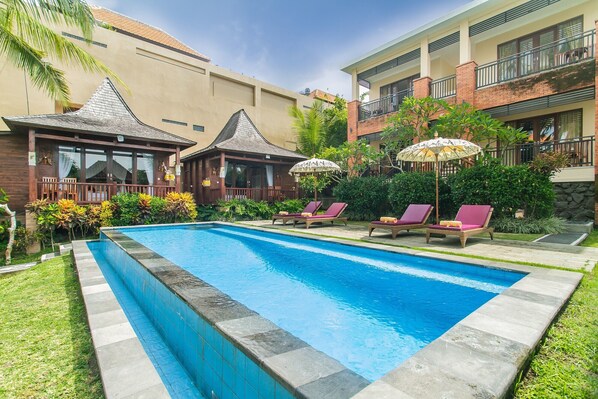 A swimming pool with lounge chairs and umbrellas, surrounded by greenery and a building with multiple balconies. - Buah Nagasari Cottages by Supala Hospitality (Ubud)