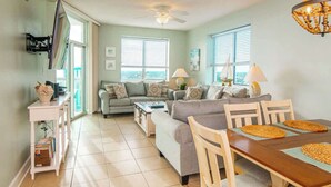 A living room with a sofa, coffee table, and dining table with chairs. There are windows with blinds, a ceiling fan, and a chandelier. - Penthouse Stunning Ocean Views Lazy River Indoor Pool (North Myrtle Beach)