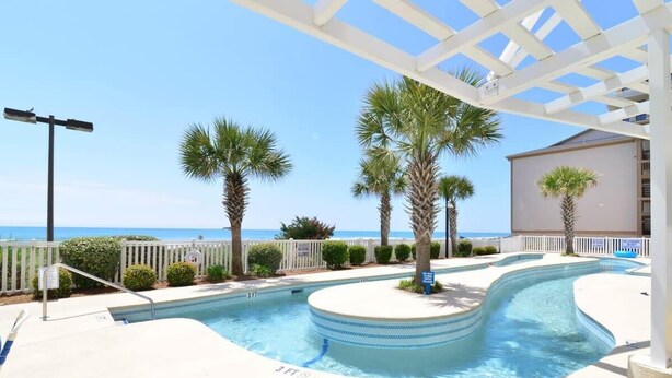 A swimming pool with a curved design, surrounded by palm trees and a white pergola. - Penthouse Stunning Ocean Views Lazy River Indoor Pool (North Myrtle Beach)