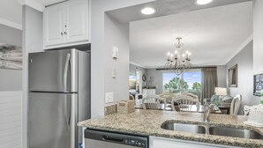 A modern kitchen with a stainless steel refrigerator, granite countertops, a dishwasher, and a dining area with a chandelier. - 🌅Islander #204 Luxury Living with Sunrise Views & Gulf Wildlife Watching🌊🐬 (Destin)