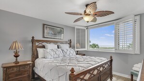 A bedroom with a wooden bed, a nightstand, a ceiling fan, a window with shutters, and a view of the outdoors. - 🌅Islander #204 Luxury Living with Sunrise Views & Gulf Wildlife Watching🌊🐬 (Destin)