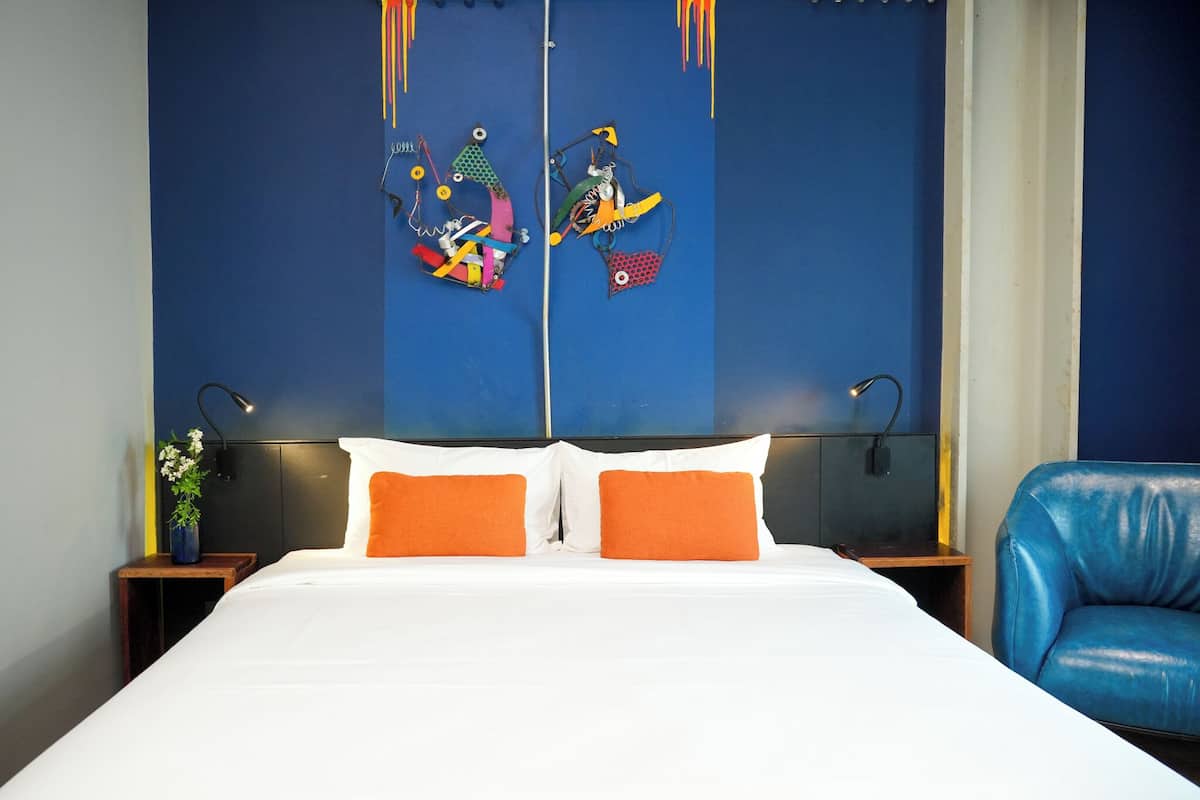 A hotel room with a large bed, two orange pillows, a blue leather chair, and a wall with colorful artwork.