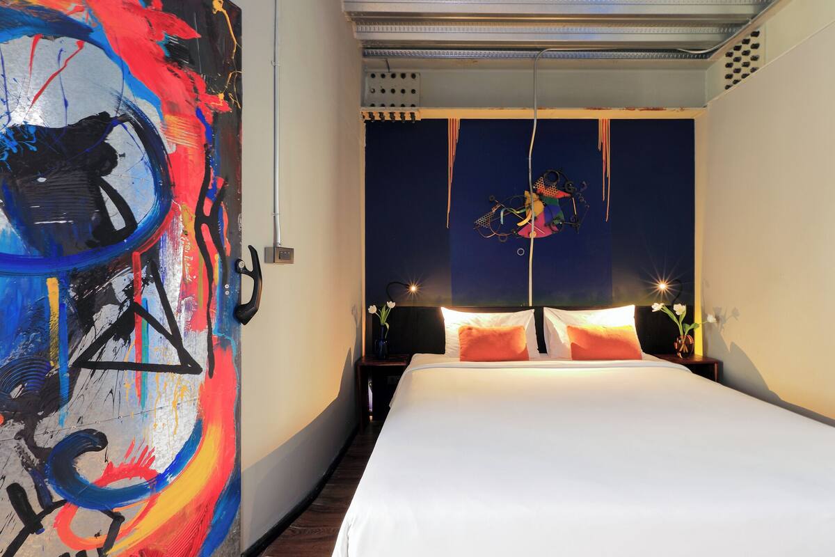 A bedroom with a large abstract painting on one wall, a bed with white bedding, and a nightstand with a vase of flowers.