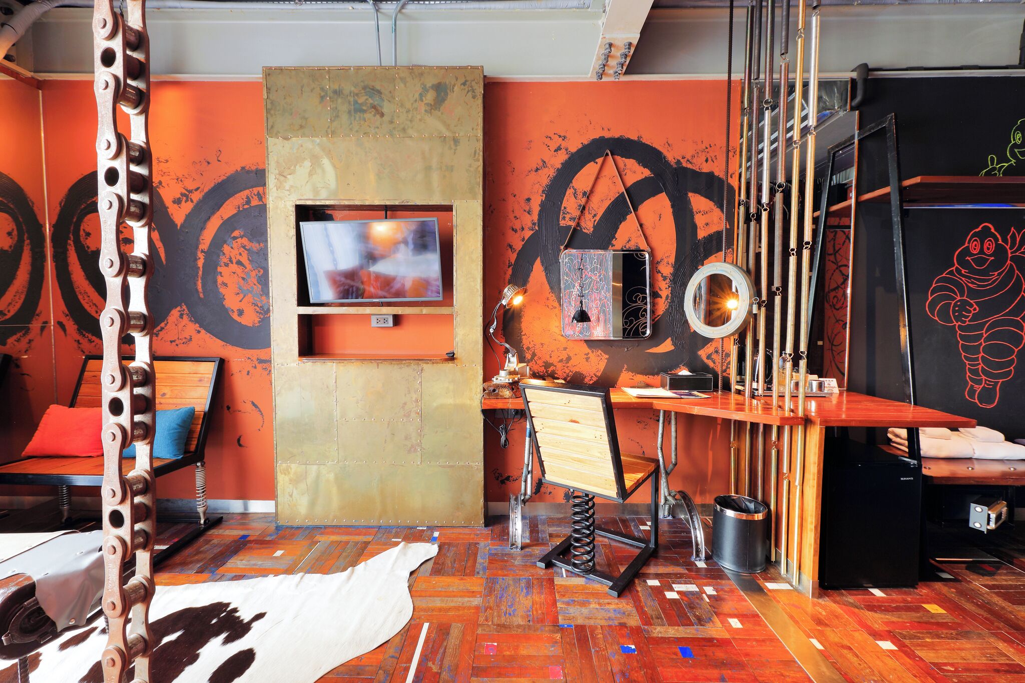 A room with a wooden floor, a bed with a red pillow, a TV mounted on a wall, a desk with a chair, and a refrigerator.