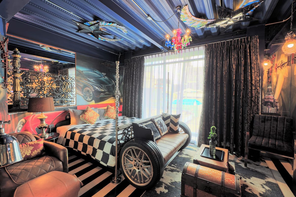 A room with a checkered bed, a sofa, a vintage lamp, and a large window with curtains.