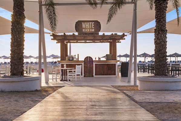 Eine Strandbar namens "WHITE BEACH BAR" mit einer Holzpergola, Sitzgelegenheiten und Palmen.