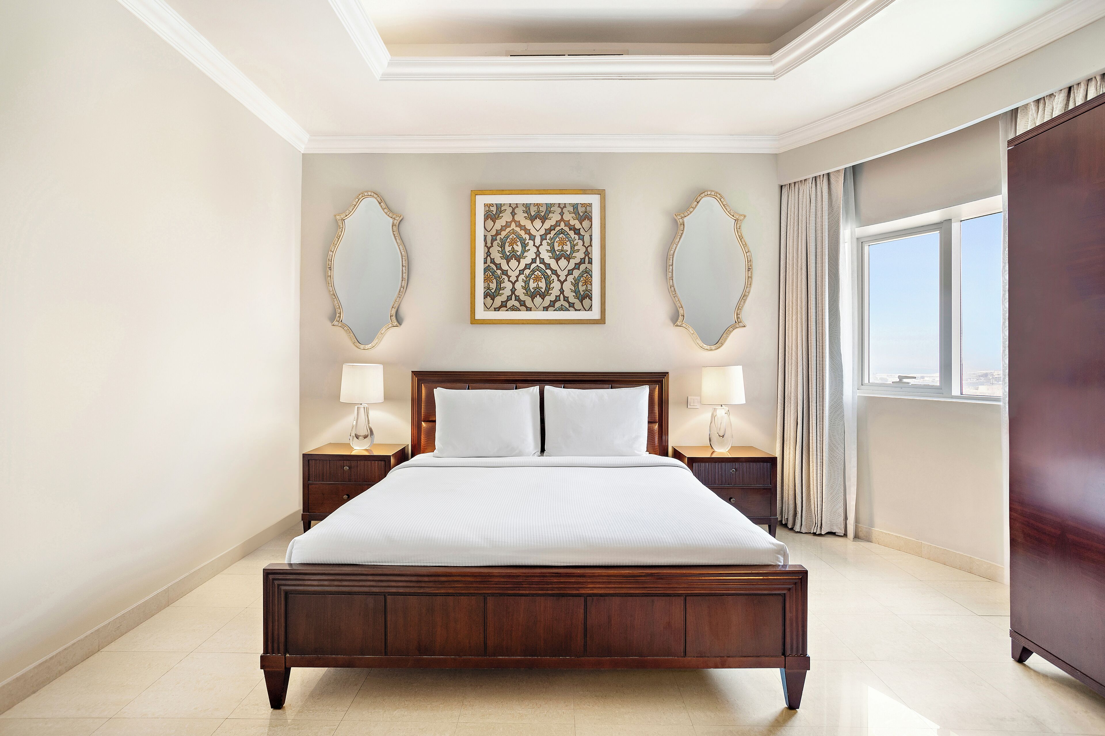 A large bed with a wooden frame, two nightstands with lamps, a decorative mirror, and a window with curtains.