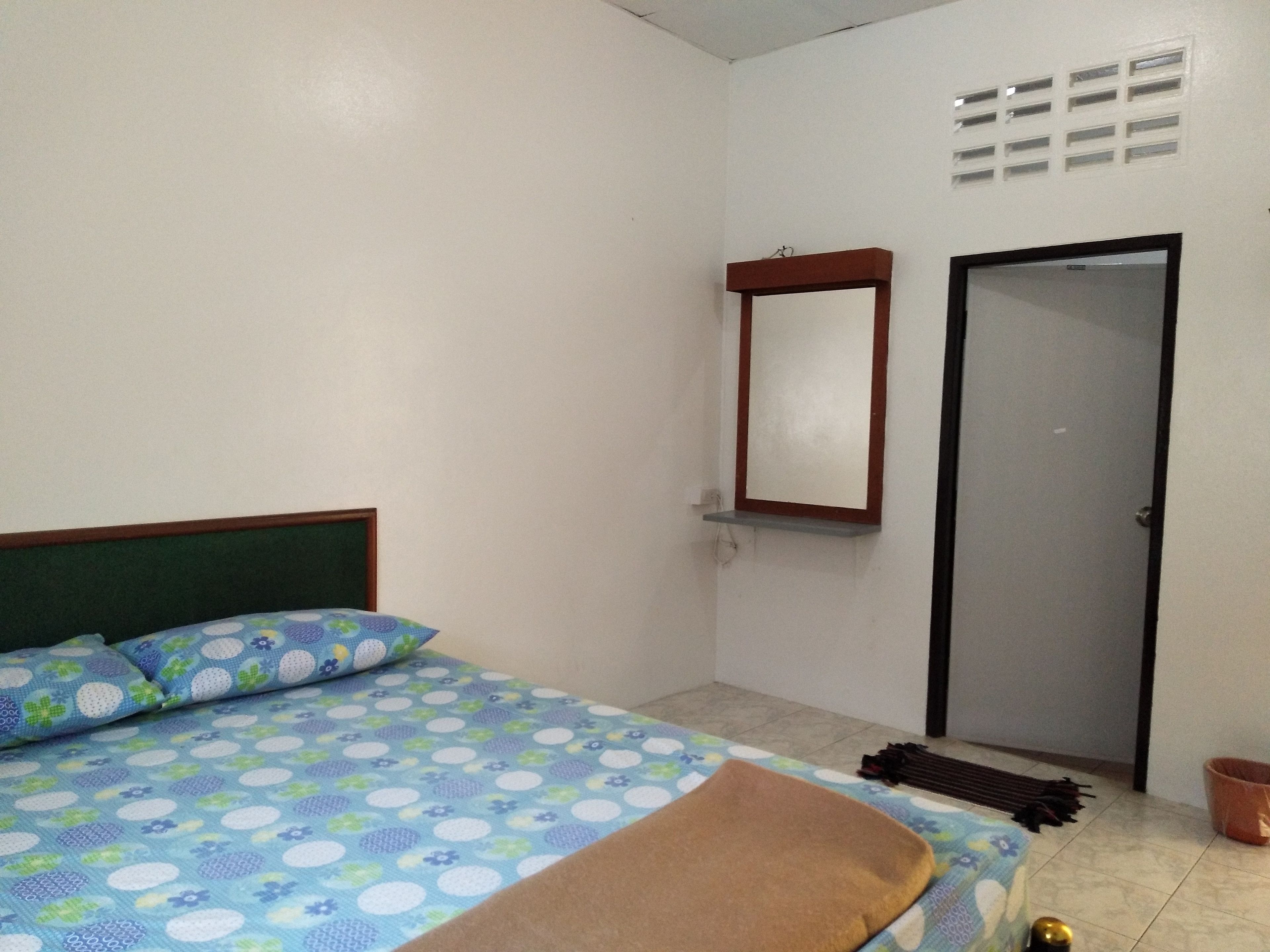 A bedroom with a bed, a bedside table, a mirror, a door, and a window with a vent.