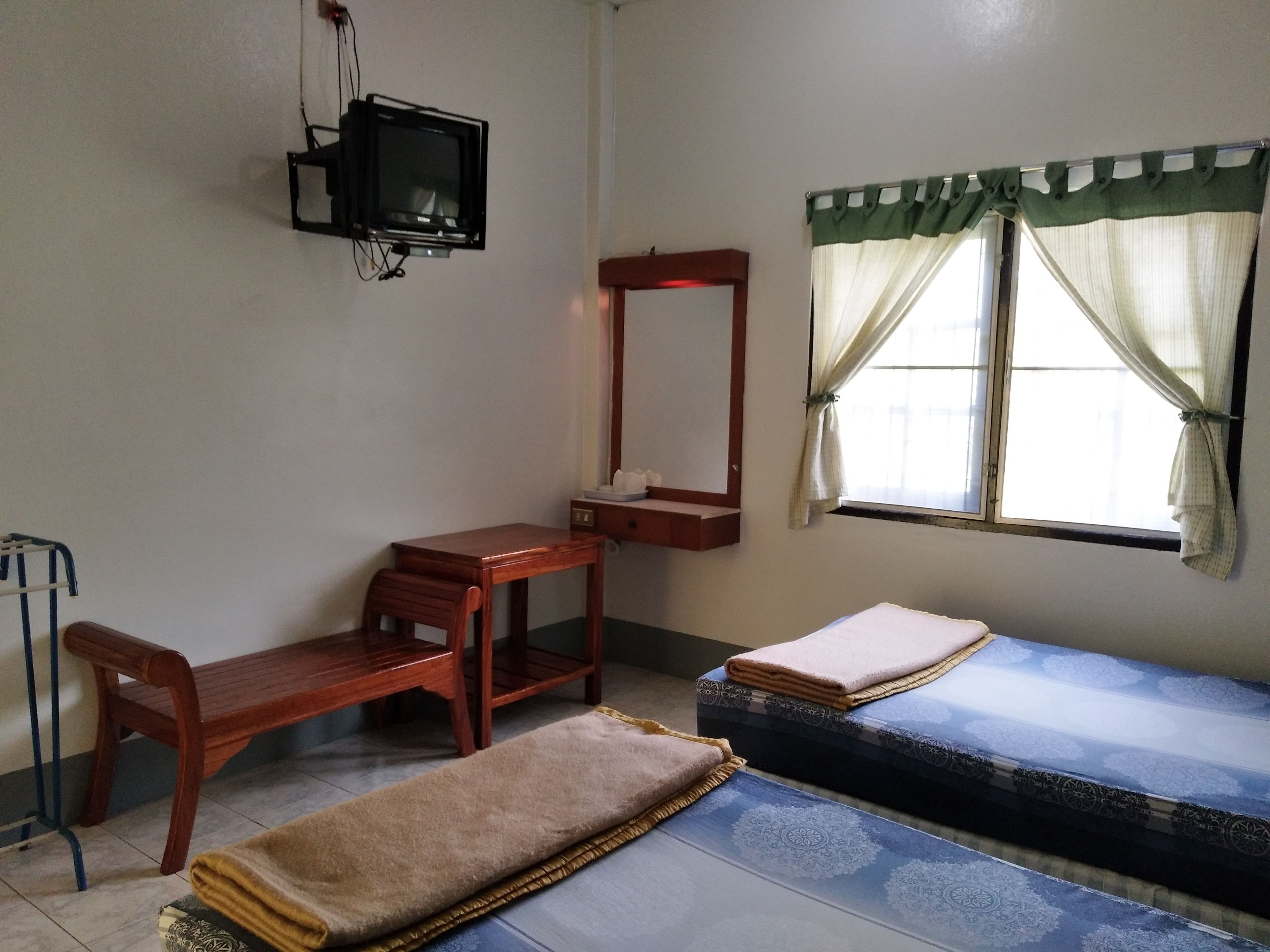 Una habitación con escritorio de madera, una silla, una cama con colchón estampado, una mesita de noche con espejo y un televisor montado en la pared.