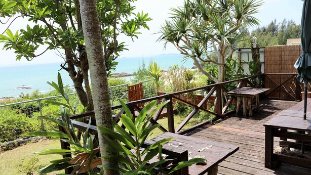 A wooden deck with tables and chairs, surrounded by tropical plants and overlooking a scenic view of the ocean. - Uminosoba Pension (Ginoza)