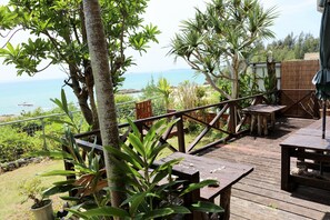 A wooden deck with tables and chairs, surrounded by tropical plants and overlooking a scenic view of the ocean. - Uminosoba Pension (Ginoza)