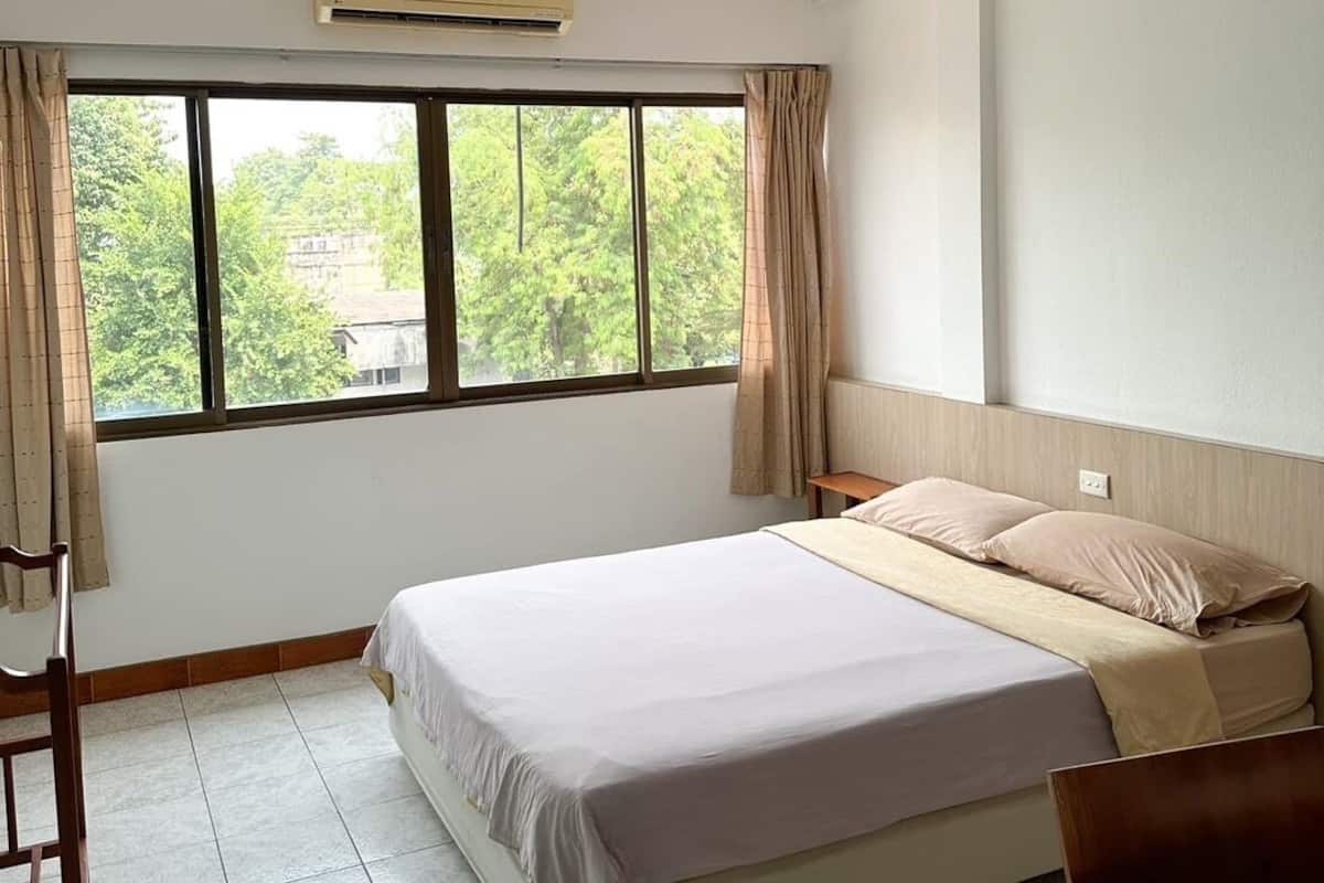 Une chambre à coucher avec un lit, une fenêtre, un appareil de climatisation et une vue sur les arbres et les bâtiments environnants.