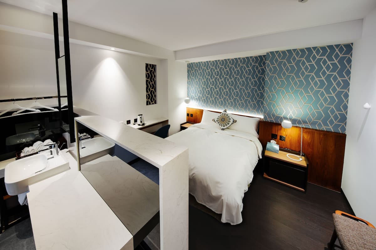 A modern hotel room with a bed, bedside tables, a desk, and a bathroom with a sink and mirror.