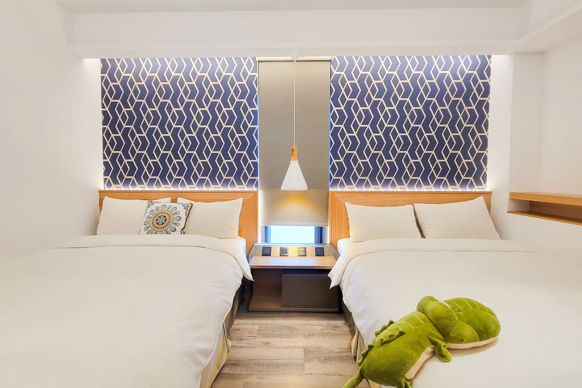 A hotel room with two beds, a nightstand, a lamp, and a green plush toy on the right bed.