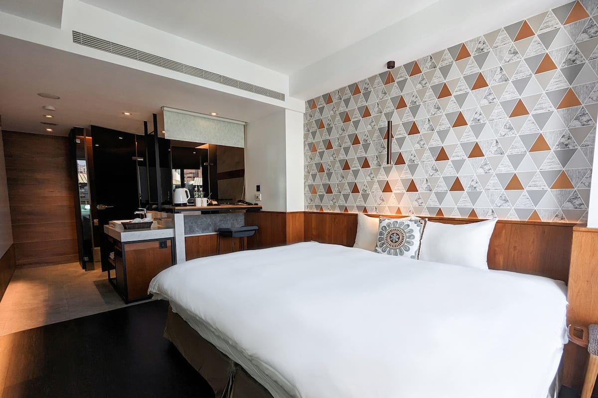 A modern hotel room with a large bed, a built-in desk with a coffee maker, and a bathroom with a mirror and sink.