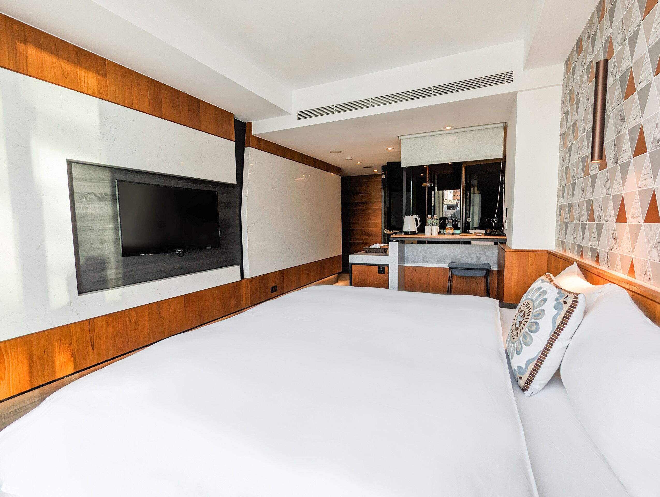 A modern hotel room with a large bed, a TV mounted on the wall, a small kitchenette, and a bathroom with a mirror.