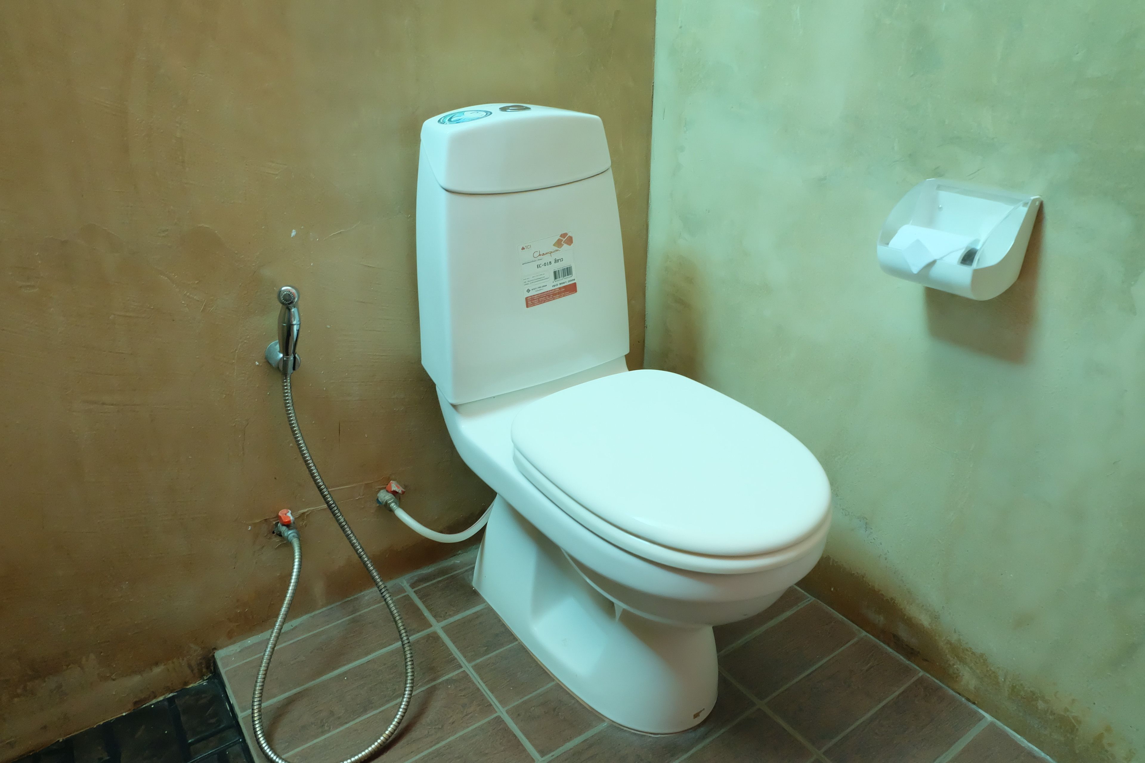 Des toilettes blanches, avec un réservoir et un accessoire de type bidet, dans une salle de bain dont le sol est carrelé et dont un des murs est uni.