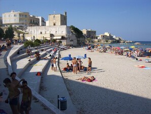 Une plage où des gens se prélassent au soleil et se baignent, un escalier menant à l’eau, et des bâtiments en arrière-plan.