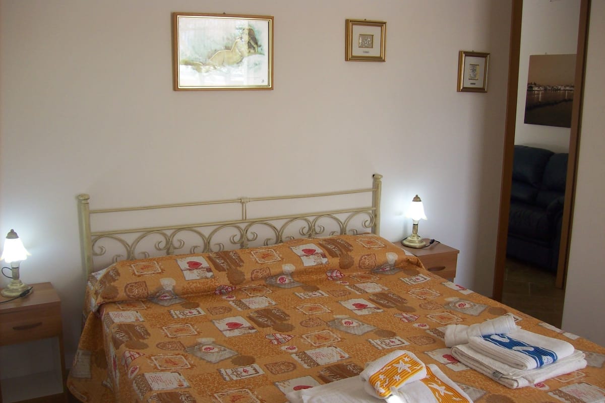 Ein ordentlich bezogenes Bett mit einem gemusterten Überwurf, zwei Nachttischen mit Lampen und einem gerahmten Bild an der Wand.
