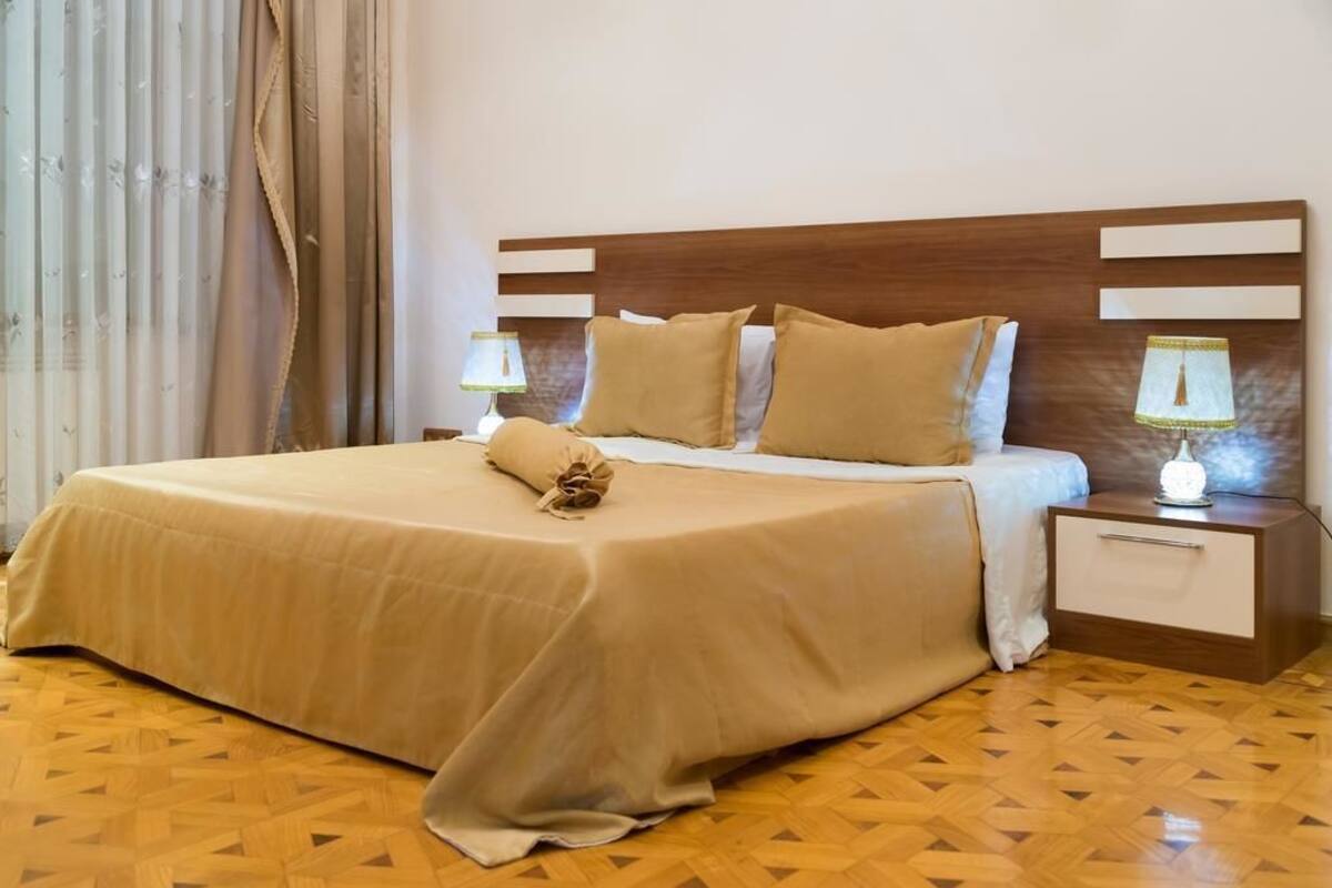 Un lit bien fait, recouvert d’une couvre-lit beige, avec deux oreillers et un objet décoratif posé dessus. Deux tables de chevet avec des lampes se trouvent de part et d’autre du lit. La pièce est dotée d’un plancher en bois avec un motif géométrique.