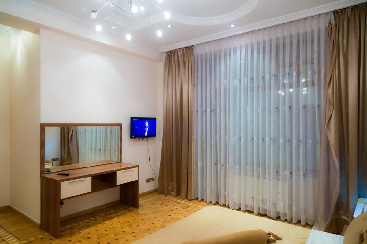 Une chambre avec un bureau en bois, une télévision, un lit et une grande fenêtre ornée de rideaux légers.