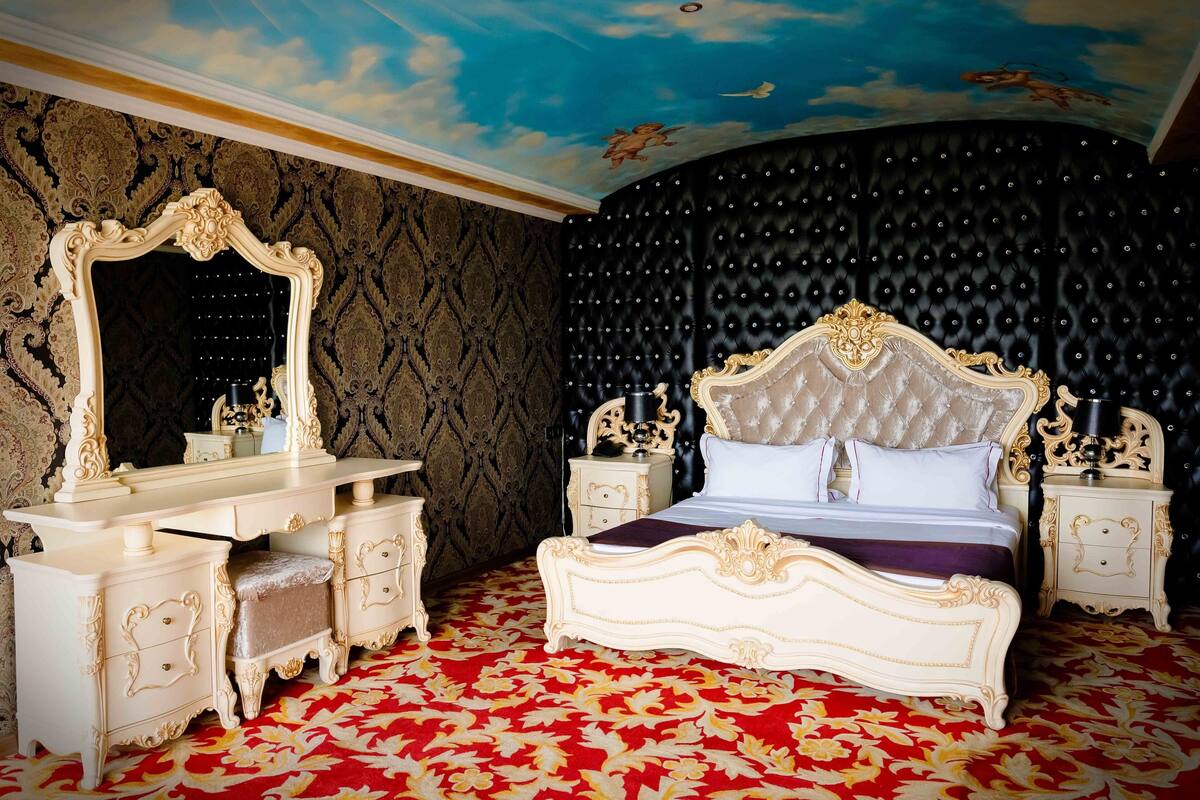 Une chambre à coucher luxueuse, dotée d’un grand lit à baldaquin richement décoré, d’une coiffeuse assortie et d’une fresque peinte au plafond.