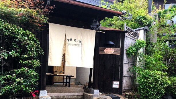 Ein traditionelles japanisches Gebäude mit Holzfassade, Steinstufen, die zu einer Holztür führen, und einer mit japanischen Schriftzeichen versehenen Fahne.