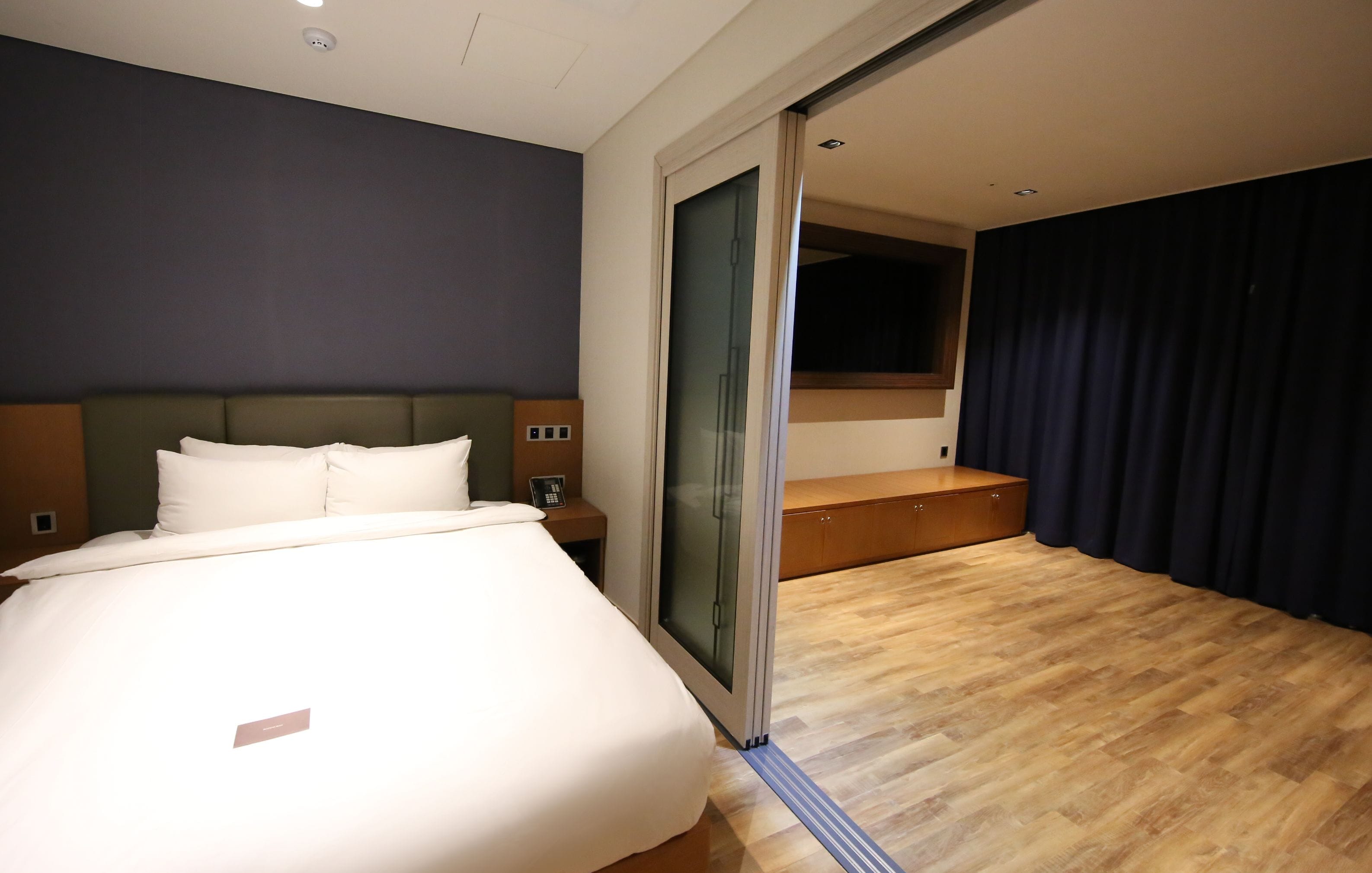 A modern hotel room with a large bed, a bedside table with a phone, a sliding glass door leading to a balcony, and a wooden floor.