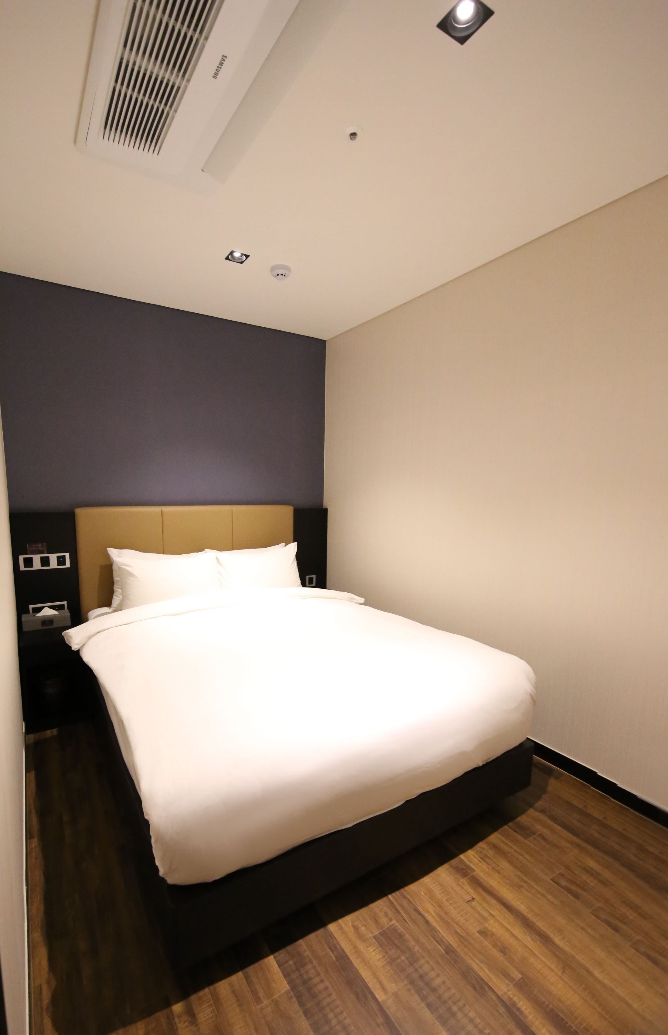 A neatly made bed in a hotel room with a dark headboard, a nightstand with a lamp, and a ceiling vent.