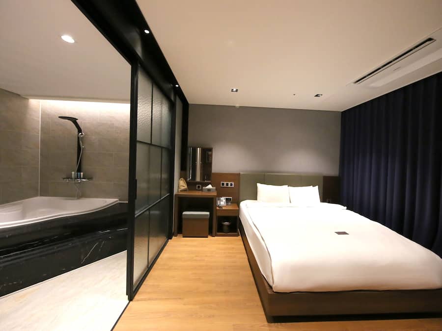 Una habitación de hotel moderna con una bañera grande, una cama, una mesita de noche y un espejo.
