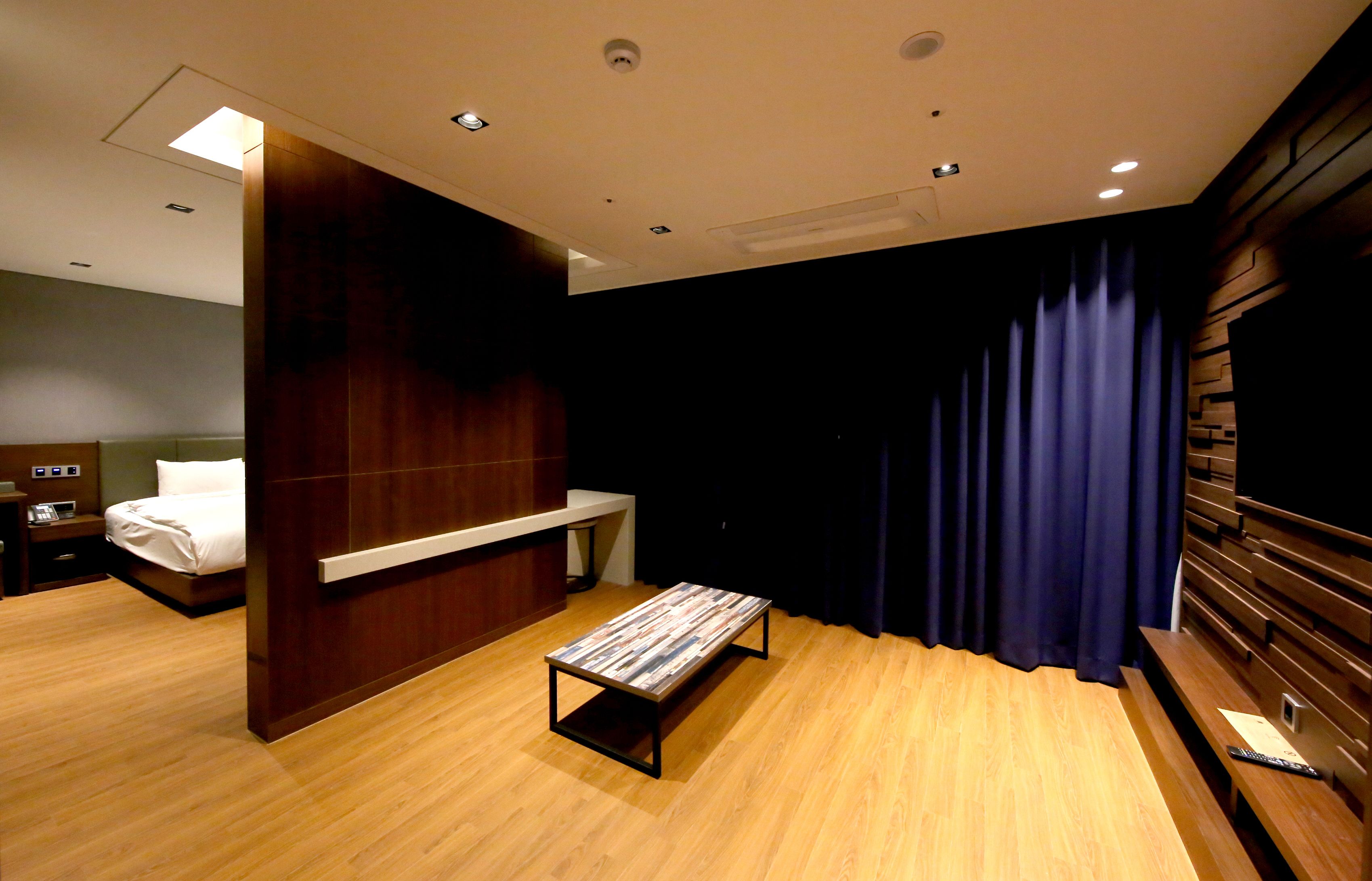 A modern hotel room with a wooden floor, a bed with white linens, a small table, and a dark curtain.
