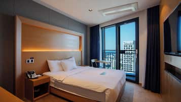 Habitación de hotel con una cama grande, una mesita de noche con un teléfono, una pequeña mesa con una silla y vistas a un paisaje urbano a través de un ventanal amplio.