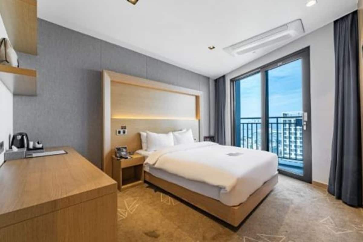 A hotel room with a large bed, a desk, a nightstand, and a view of the city through large windows.