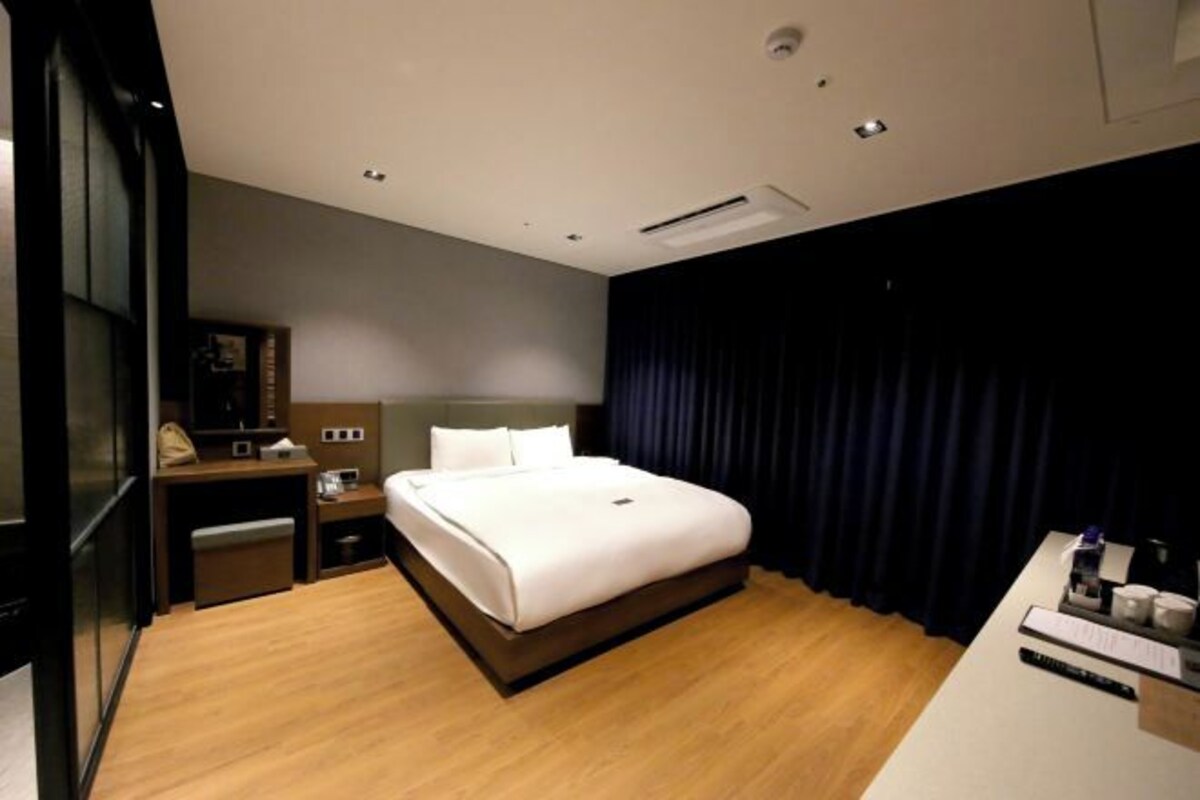 A hotel room with a large bed, a desk with a chair, a desk lamp, a telephone, and a dark curtain.