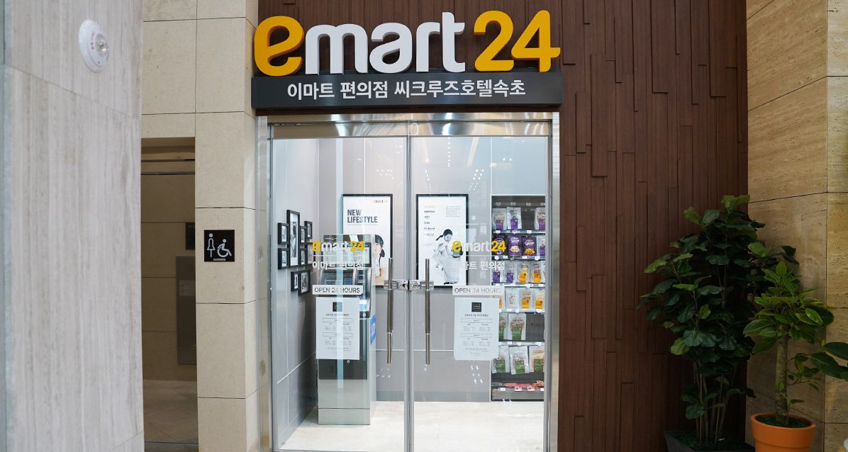 The entrance to emart 24, a 24-hour convenience store with glass doors, a wheelchair accessibility sign, and a potted plant to the right.