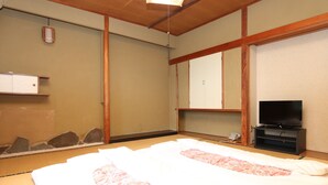 Una habitación tradicional japonesa con suelo de tatami, una cama con colcha floral, un televisor sobre un soporte y un armario de madera.