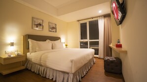 Habitación de hotel con una cama grande, mesitas de noche, un taburete pequeño, una televisión montada en la pared y una ventana con cortinas.