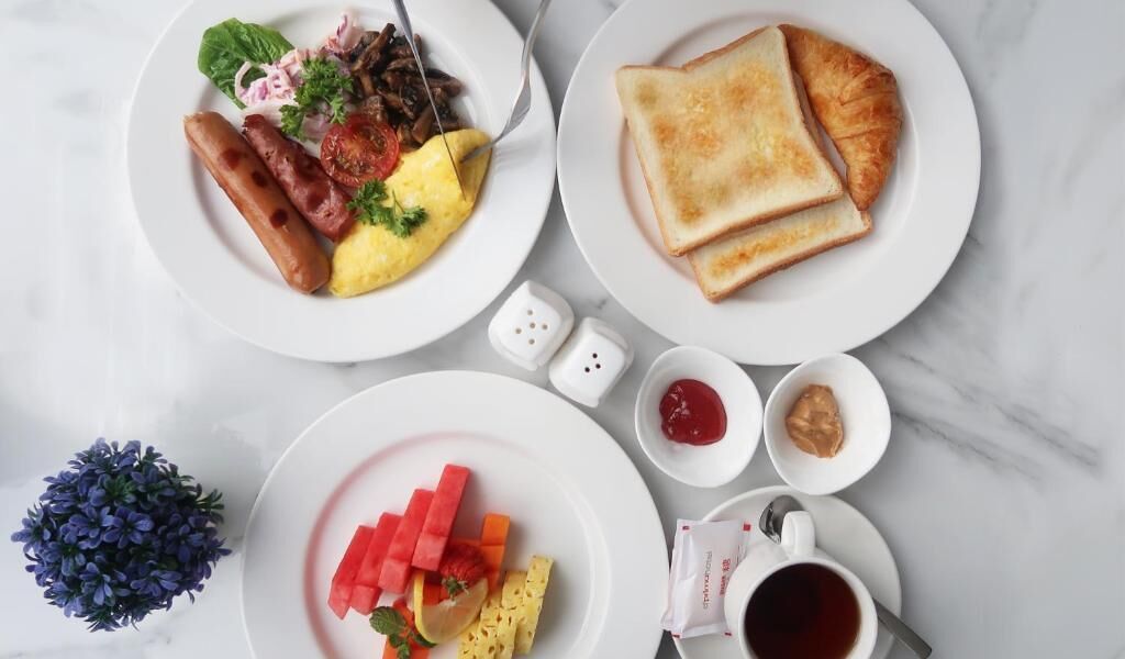 Ein Frühstückstisch reichhaltig gedeckt mit Speisen, darunter Würstchen, Eier, Toast und Obst, sowie Soßen und einer Kaffeetasse.