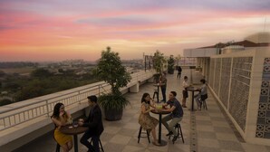 Terraza en la azotea con varias mesas y sillas, rodeada por una barandilla y vistas a la ciudad de fondo.