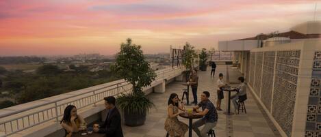 A rooftop terrace with multiple tables and chairs, surrounded by a railing, and a view of the cityscape in the background.