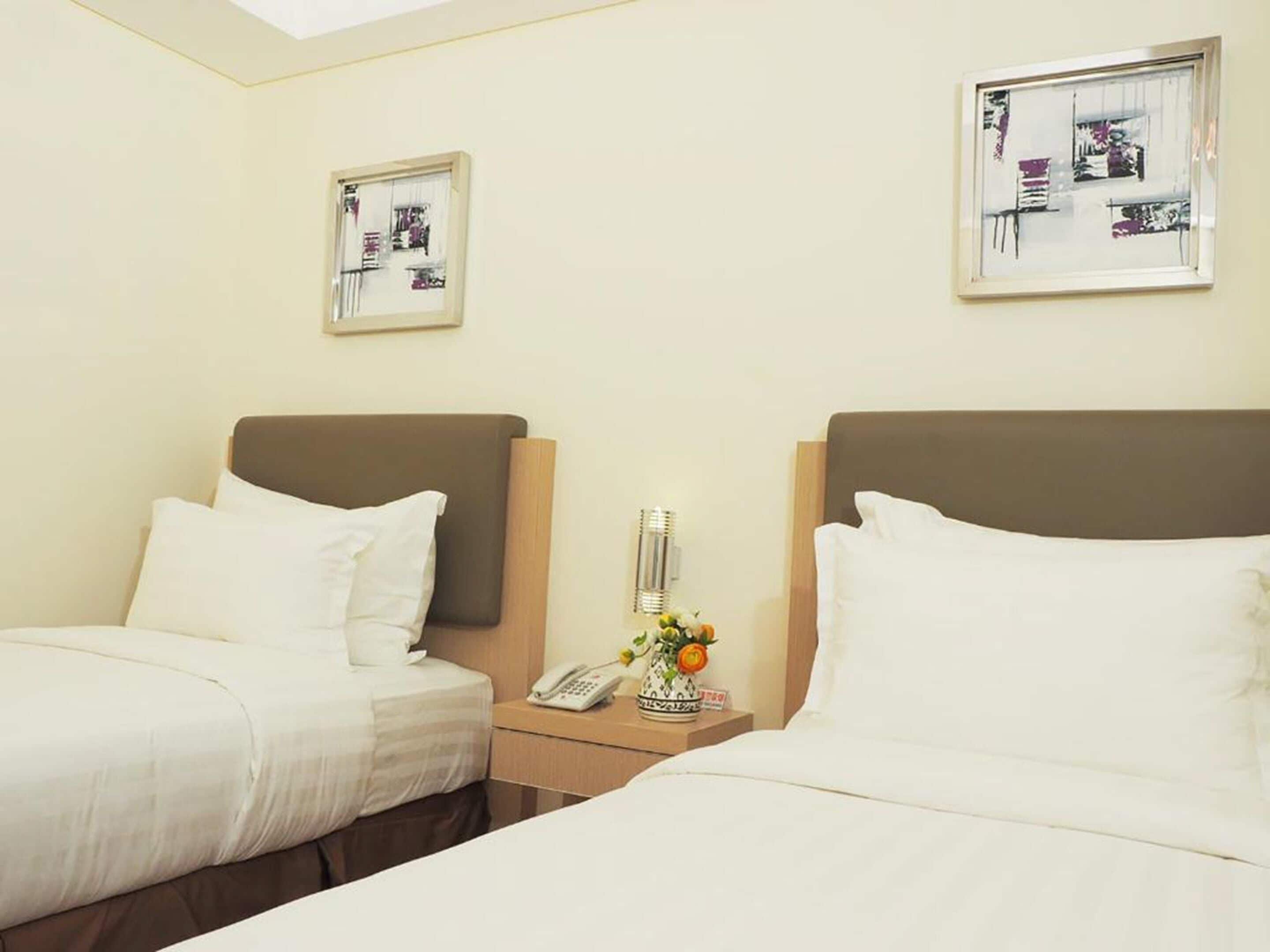 Ein Hotelzimmer mit zwei Betten, einem Nachttisch mit Telefon und Blumenvase sowie zwei gerahmten Bildern an der Wand.