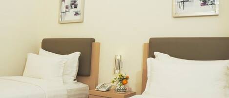 Ein Hotelzimmer mit zwei Betten, einem Nachttisch mit Telefon und Blumenvase sowie zwei gerahmten Bildern an der Wand.