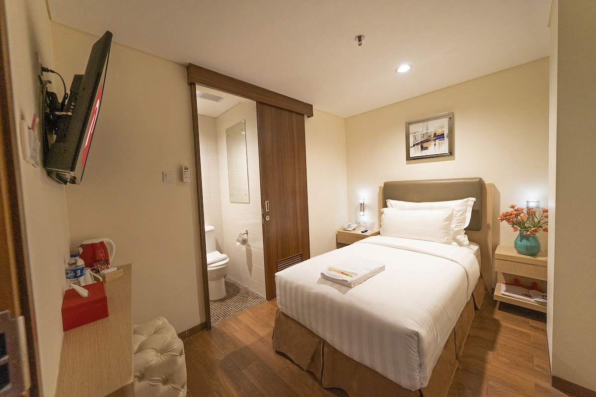 Habitación de hotel con cama, mesita de noche, televisor montado en la pared, baño visible a través de una puerta abierta y un cuadro enmarcado en la pared.