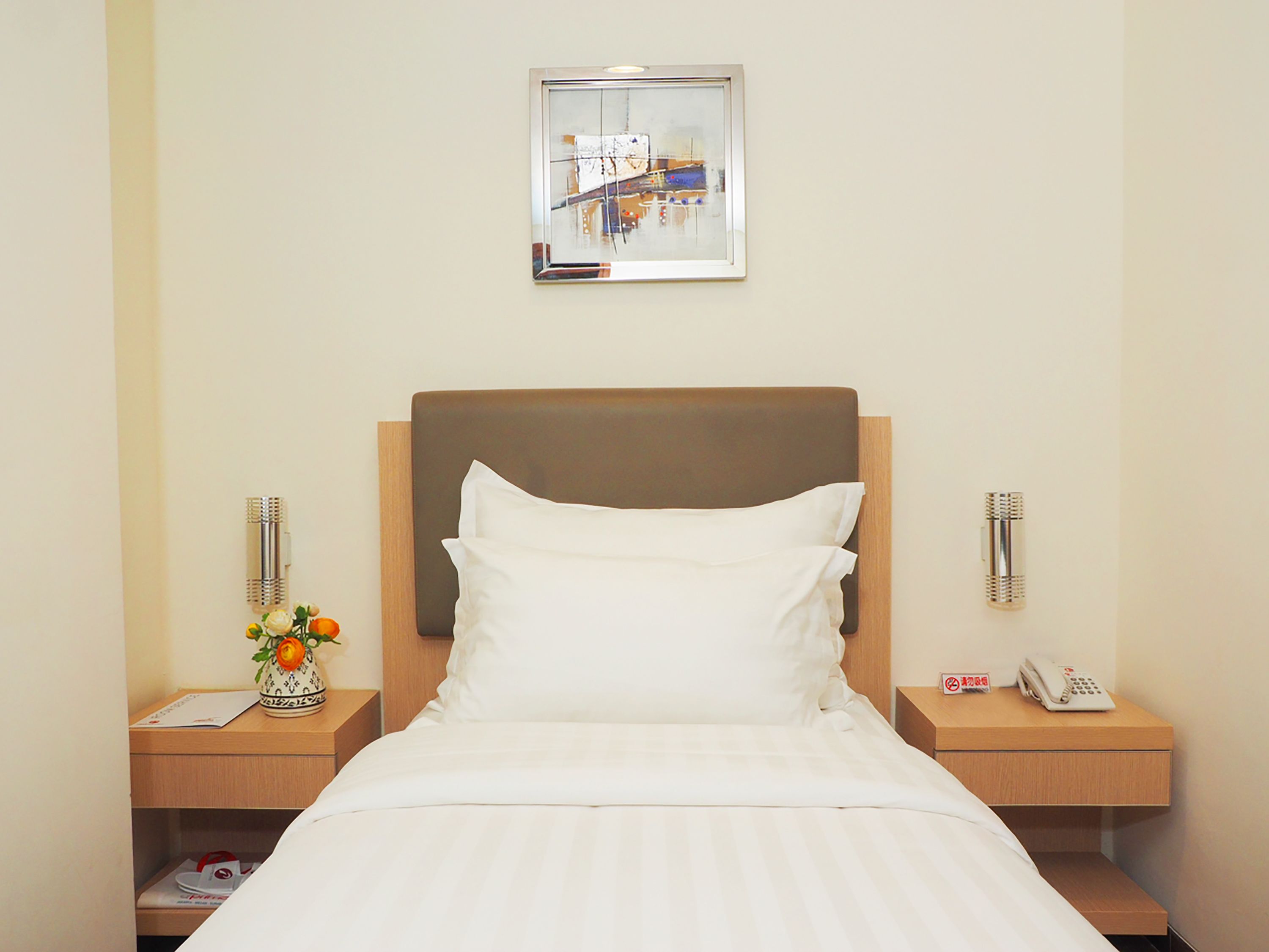 A hotel room with a bed, two bedside tables, a nightstand, a framed picture on the wall, and a vase with flowers.