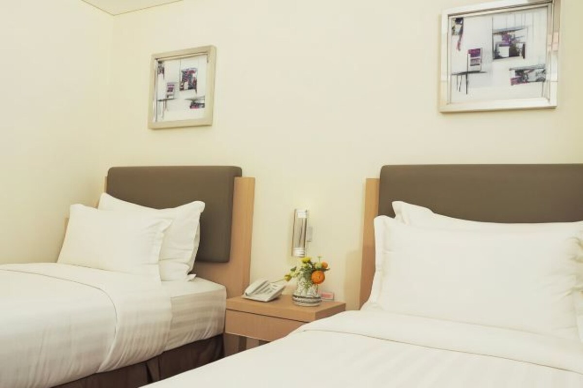 Quarto de hotel com duas camas, uma mesa de cabeceira com telefone e um vaso de flores, e quadros nas paredes.