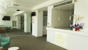 A modern reception area with a glass partition, a reception desk with a logo, and a seating area with teal chairs. - Hotel Sol de Jávea (Javea)