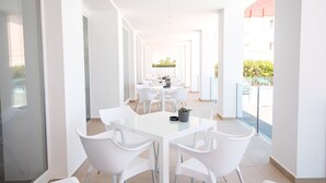 A bright, modern dining area with white tables and chairs, a potted plant, and a view of the outside. - Hotel Sol de Jávea (Javea)